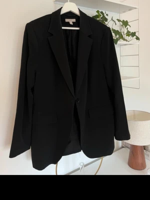 Svart klassisk kavaj med knappar - Stilren svart kavaj med klassisk passform och enkelknäppning framtill. Kavajen har två fickor och tre dekorativa knappar vid ärmsluten. Perfekt för en dressad look och passar till många olika outfits.