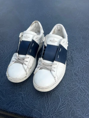Valentino vita sneakers  - Snygga vita sneakers från Valentino med bred mörkblå remsa över mitten och klassisk logga på plösen. Skorna har vita snören och är tillverkade i slätt läder. Perfekta för dig som vill ha en stilren och exklusiv look. Storlek 43 skorna är äkta hör av er vid frågor pris går att diskutera 