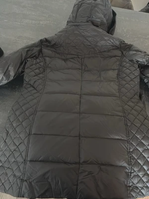 Svart dunjacka Rockandblue dam - Snygg svart dunjacka från Rockandblue med huva och quiltade detaljer på sidorna och ärmarna. Jackan har en figurnära passform och är perfekt för kalla vinterdagar. Klassisk design som funkar till det mesta och håller dig varm hela säsongen.