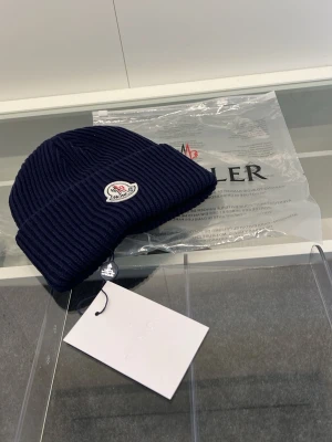 Mörkblå ribbstickad Moncler mössa - Säljer en mörkblå ribbstickad mössa från Moncler med uppvikt kant och Monclers klassiska logotyp framtill. Mössan är tillverkad i mjukt material och har en stilren, tidlös design som passar perfekt till kalla dagar. Obs vid köp välj PostNord frakt, priset kan prutas vid snabb affär. Hör av vid funderingar eller mer bilder👍