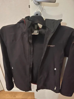 Svart vindjacka från Everest - Snygg svart vindjacka från Everest med dragkedja framtill, huva och två fickor med dragkedja. Jackan har diskret logga på bröstet och är tillverkad i ett lätt och slitstarkt material som passar perfekt för blåsiga dagar.