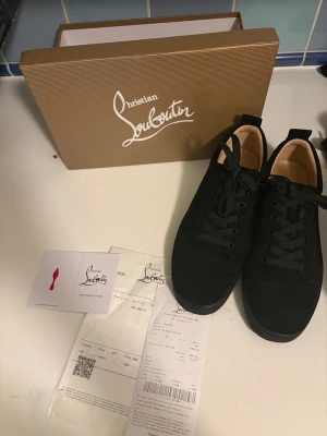 Svarta sneakers från Christian Louboutin - Stilrena svarta sneakers från Christian Louboutin med klassisk röd sula och svarta skosnören. Ovandelen är i mocka och insidan har en beige ton. Kommer med originalkartong och tillbehör. Perfekta för dig som vill ha en lyxig och tidlös sneaker.