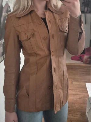 Brun vintage skinnjacka med fickor - Trendig brun skinnjacka med markerad midja och stora kragar. Jackan har två bröstfickor med lock och knapp, samt knäppning framtill med matchande knappar. Långa ärmar och tydliga sömmar ger en cool vintagekänsla. Perfekt att styla till jeans eller kjol. Storlek 38 men är mer som en 36. Jag har storlek 34 i vanliga fall, se bilder hur den sitter på mig.