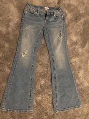 Ljusblå bootcut jeans med slitningar - Säljer ett par ljusblå bootcut jeans med snygga slitningar på låren. Jeansen har låg midja och är tillverkade i mjuk denim med stretch. Perfekta för en avslappnad och trendig look.