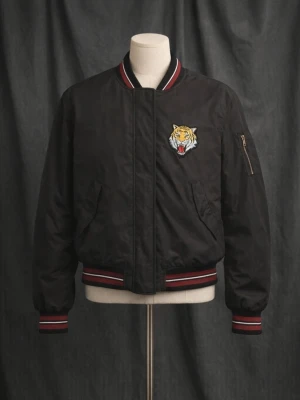 Svart bomberjacka med tigerpatch - Cool svart bomberjacka med broderad tigerpatch på bröstet. Jackan har ribbade muddar med röda och vita ränder, dragkedja och fickor samt en ärmficka med zip. Perfekt för dig som vill sticka ut med en edgy look.