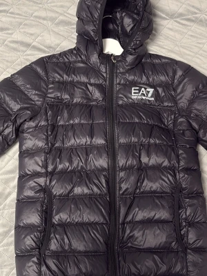 Äkta svart dunjacka från EA7 Emporio Armani - Snygg svart dunjacka från EA7 Emporio Armani med huva och dragkedja framtill. Köpte för 1500 kr, men säljer för 550. Ja, den är äkta. Kvalitet 10/10, inte en endaste skråma överhuvudtaget. Säljer för att den är för liten för mig  