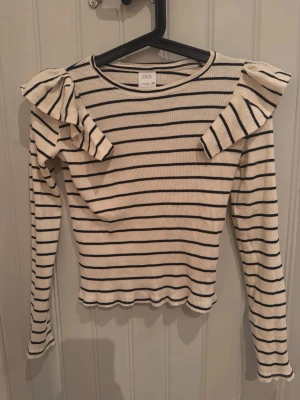 Randig topp med volang från Zara - Snygg ribbad topp från Zara med svarta ränder på beige botten. Långärmad modell med volangdetaljer på axlarna och rund halsringning. Perfekt för dig som gillar en trendig och lekfull stil.