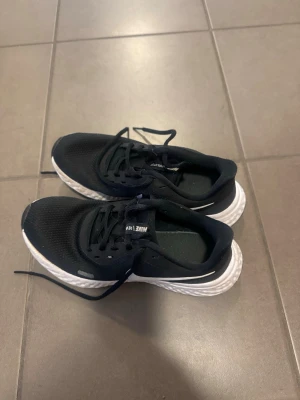 Svarta Nike sneakers med vit sula - Säljer ett par svarta Nike sneakers med vit, strukturerad sula och klassisk swoosh på sidan. Ovandelen är i mesh för bra andningsförmåga och skorna har svarta skosnören. Perfekta för dig som gillar sportig och stilren look.