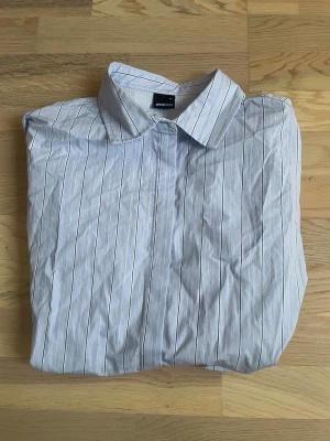 Randig skjorta från Gina Tricot  - Säljer en klassisk ljusblå skjorta från Gina Tricot med tunna vita och mörka ränder. Skjortan har krage, knappar framtill och långärmad design. Knappar i midjan för en figursydd look. I princip helt nyköpt. 