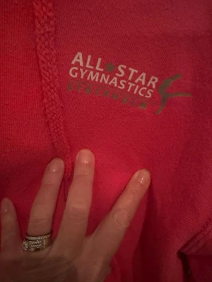 Rosa hoodie All Star Gymnastics - Säljer en rosa hoodie från All Star Gymnastics Stockholm. Hoodien har huva med dragsko, stor magficka och tryck på bröstet. Perfekt för dig som gillar gymnastik och vill ha en skön och sportig stil. Materialet känns mjukt och bekvämt.