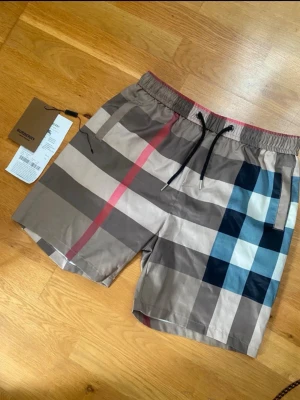 Burberry shorts - Säljer ett par klassiska rutiga shorts från Burberry med beige, svart, blå och röda toner. Shortsen har elastisk midja med snörning och sidofickor. De är oanvända och i mycket fint skick. Perfekta för sommaren och ger en stilren vibe till din outfit.    Pris kan diskuteras vid snabb affär.