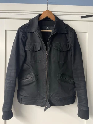 Svart jeansjacka från Tiger of Sweden - Stilren svart jeansjacka från Tiger of Sweden med dragkedja framtill, två bröstfickor med lock och två snedställda fickor med dragkedja. Klassisk krage och raka ärmar ger en modern look. Perfekt för dig som gillar minimalistisk stil.