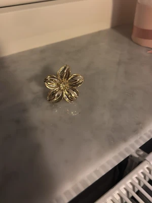 Guldfärgad blomring i metall - Snygg ring i guldfärgad metall formad som en stor blomma med fem blad. Ringen har en glansig finish och detaljer i mitten som ger extra bling. Perfekt för dig som vill sticka ut med ett unikt smycke. Från new yorker. Storlek s
