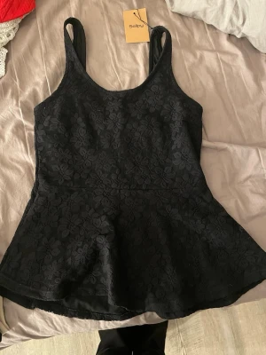 Svart blommig linnetopp i spets - Snygg svart linnetopp med blommigt spetsmönster och breda axelband. Toppen har en lätt utsvängd form nedtill och är perfekt för dig som gillar en feminin och trendig look. Passar till både jeans och kjol.