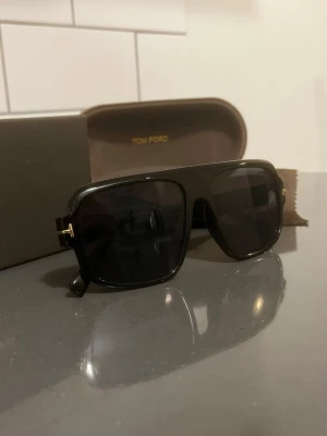 Svarta solglasögon från Tom Ford - Snygga svarta solglasögon från Tom Ford med breda bågar och mörka glas. Klassisk och stilren design med guldfärgade detaljer vid gångjärnen. Kommer med originalfodral och putsduk. Perfekt för dig som vill ha en cool och exklusiv look.