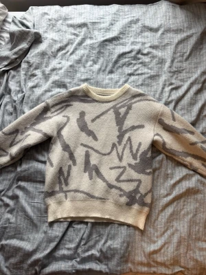Beige stickad tröja med grått mönster - Säljer en beige stickad tröja med abstrakt grått mönster. Tröjan har rund halsringning och ribbstickade muddar vid ärmslut och nederkant. Perfekt för dig som gillar en unik och trendig look.