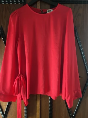Röd långärmad blus med knytband - Trendig röd blus från Indiska med rund halsringning och långa ärmar. Blusen har ett snyggt knytband i sidan och lätt transparent material som ger en luftig känsla. Perfekt för dig som vill ha en färgstark och stilren look.
