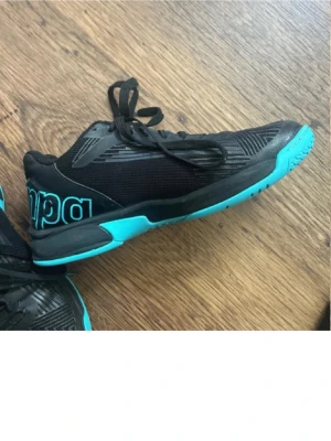 Svarta sneakers från Kempa med turkosa detaljer - Säljer ett par svarta sneakers från Kempa med turkosa detaljer och sula. Skorna har meshpaneler för bra ventilation och stora logotyper på sidorna. Perfekta för dig som gillar sportig stil och vill sticka ut med färg. Använd som handboll/inneskor i ca 6 månader, otroligt sköna