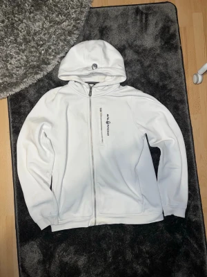 Vit hoodie från Sail Racing - Säljer en vit hoodie från Sail Racing med dragkedja framtill och en bröstficka med zip. Logga på bröstet och på huvan. Hoodien har en sportig look och är tillverkad i mjukt material, perfekt för chill dagar eller när du vill ha en clean stil.lite söndersliten vid hönderna annars inga problem ! 