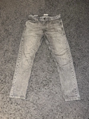 Jack & Jones - Säljer ett par grå tvättade jeans från Jack & Jones med rak passform och klassisk femficksdesign. Jeansen har en snygg vintagekänsla och är tillverkade i denim med tydlig tvättad effekt. Perfekta för en avslappnad och trendig look.