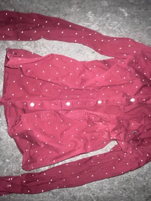 Röd prickig skjorta med knappar - Snygg röd skjorta med vita prickar och klassisk knappstängning framtill. Skjortan har lång ärm och är tillverkad i ett mjukt bomullstyg som känns skönt mot huden. Perfekt för dig som vill sticka ut lite extra med ett lekfullt mönster.