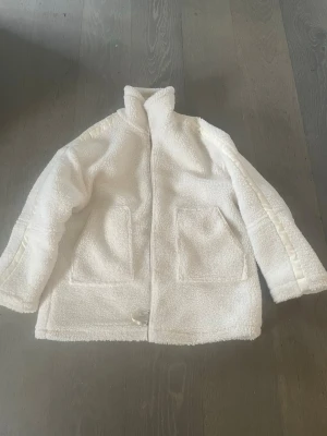 Vit teddyjacka med hög krage - Mysig vit teddyjacka med hög krage och två stora framfickor. Jackan har dragkedja framtill och detaljer i samma färg längs axlarna. Perfekt för kalla dagar och ger en trendig look med sin fluffiga struktur.