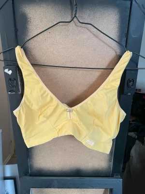 Gul bikiniöverdel från Gina Benotti - Snygg gul bikiniöverdel från Gina Benotti med breda axelband och dekorativt GB-hänge framtill.