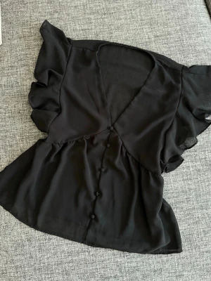 Svart blus med volangärmar - Snygg svart blus i tunn, luftig chiffong med djup V-ringning och dekorativa klädda knappar framtill. Blusen har korta volangärmar och lätt utsvängd passform som ger en feminin siluett. Perfekt att styla med jeans eller kjol.