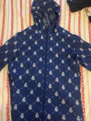 Blå vindjacka med Real Madrid-tryck - Snygg blå vindjacka med heltryck av Real Madrids klubbmärke och vita detaljer. Jackan har huva och dragkedja framtill, perfekt för dig som vill visa ditt lagstöd. Tillverkad i lätt och vindtätt material som passar till streetwear.