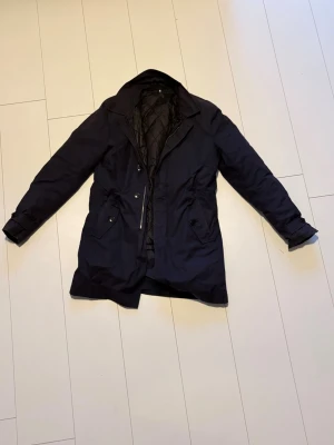 Marinblå rock från J.Lindeberg - Stilren marinblå rock från J.Lindeberg med quiltat innerfoder och två stora fickor framtill. Klassisk krage och diskret logga på ärmen. Perfekt för kyligare dagar och ger en clean look till din outfit.