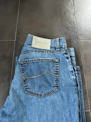 Blå jeans från Jacob Cohen - Säljer ett par klassiska blå jeans från Jacob Cohen med fem fickor och snygga kontrastsömmar. Jeansen har en ljus tvätt och patch med logga bak i midjan. Tillverkade i mjuk denim som ger en avslappnad look.