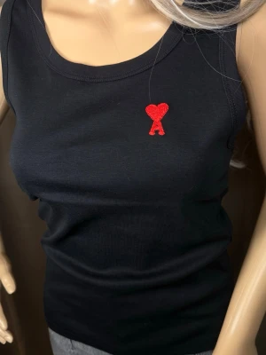 Svart linnetopp från Ami Paris - Snygg svart linnetopp från Ami Paris med det ikoniska röda hjärtat och A-broderiet på bröstet. Toppen är ribbad och har en klassisk rund halsringning. Perfekt att styla till jeans eller kjol för en trendig look.