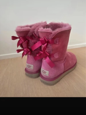 Rosa UGG boots med rosetter - Säljer ett par fluffiga rosa UGG boots med dekorativa rosetter bak. Perfekta för dig som vill ha både värme och stil. UGG-logga bak på hälen och snygga detaljer i satinband.
