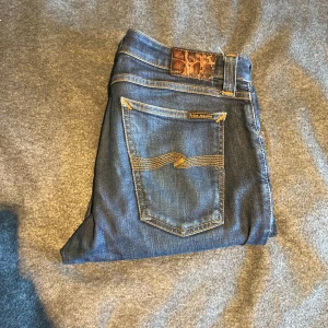 Nudie Jeans  - Säljer ett par mörkblå Nudie Jeans Skinny Lin med klassiska orange sömmar och broderade bakfickor. Jeansen har en tight passform och är tillverkade i stretchig bomullsdenim för extra komfort. Perfekta för dig som gillar en smal siluett och snygga detaljer.