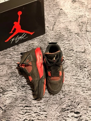 Air Jordan 4 Retro Svart/Röd - Säljer ett par Air Jordan 4 Retro sneakers i svart med röda detaljer. Skorna har meshpaneler, klassisk snörning och synlig Air-enhet i sulan. Ikonisk Jumpman-logga på plösen och yttersulan. Perfekta för dig som gillar streetwear och sportig stil.