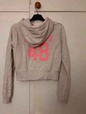 Ljusgrå hoodie med dragkedja - Ljusgrå hoodie med dragkedja framtill och två fickor. På ryggen står det 'New York 48' i neonrosa tryck. Köpt här på plick  men nu är det dags för en ny ägare som ska få bära denna. Supermjuk och go