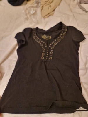 Svart topp med nitar från KappAhl - Svart kortärmad topp från KappAhl Casual Collection med dekorativa silverfärgade nitar längs v-ringningen. T-shirten har en figurnära passform och är tillverkad i mjuk bomull, perfekt för en trendig och avslappnad look.