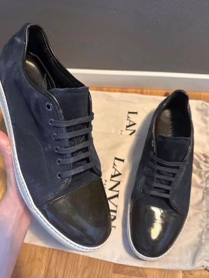 Svarta/mörkblå Lanvin sneakers med lackdetalj - Snygga svarta sneakers från Lanvin med ovandel i mocka och tåparti i blankt lack. Skorna har svarta skosnören och vit sula som ger en clean kontrast. Perfekta för dig som gillar stilrena och exklusiva sneakers med unika materialval. Skobox inte ej.          Uk storlek 15 vilket motsvarar stl 49