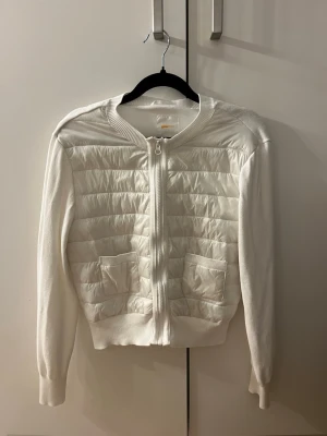 Vit quiltad cardigan med dragkedja - Stilren vit cardiganjacka med quiltade partier framtill och två fickor. Jackan har dragkedja, ribbade muddar och rund hals. Materialmix med stickade ärmar och quiltad front ger en modern look.