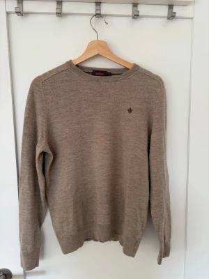 Beige stickad tröja från Morris - Klassisk beige stickad tröja från Morris med rund halsringning och diskret broderad logga på bröstet. Tröjan har långa ärmar och ribbade muddar vid ärmslut och nederkant. Perfekt basplagg i mjukt material. Tröjan har krympt i tvätten skulle därför påstå att den ligger mellan M och S