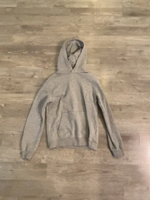 Grå hoodie med huva - En enkel och stilren grå hoodie från H&M med klassisk huva och magficka. Tillverkad i mjukt material som passar perfekt för en avslappnad look. Ribbstickade muddar vid ärmslut och nederkant ger en skön passform.
