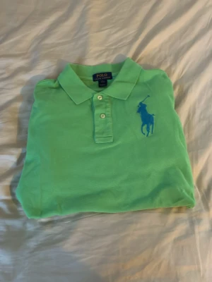 Grön piké från Polo Ralph Lauren - Snygg grön pikétröja från Polo Ralph Lauren med klassisk krage och knappar framtill. Stor blå logga broderad på bröstet och en blå detalj på ärmen. Perfekt för en avslappnad men stilren look.