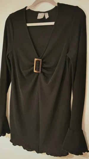 Svart v-ringad topp från ASOS - Svart långärmad topp från ASOS med v-ringning och dekorativt spänne framtill. Ärmarna har volangdetalj vid mudden och tyget känns mjukt och stretchigt. Perfekt för dig som gillar stilrena men ändå unika detaljer.