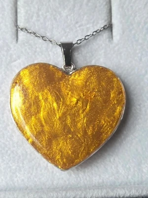 Halsband med hjärthänge i guld - Unikt halsband med ett stort hjärtformat hänge i skimrande guldton. Hänget har en metallram och sitter på en tunn silverfärgad kedja i rostfritt stål. Perfekt för dig som vill ha ett statement-smycke med glansig finish och modern känsla.