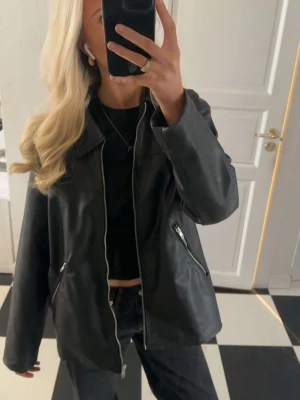 Svart oversized skinnjacka - Snygg svart skinnjacka i oversized modell med dragkedja framtill och två fickor med silverfärgade dragkedjor. Kommer inte till användning Brukar ha S o har denna i M o då sitter den lite mer löst