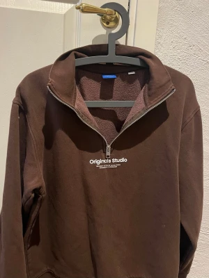 Jack & Jones brun tröja - Tröja med dragkedja framtill och texten "Originals Studio" tryckt på bröstet. Tröjan har en hög krage och långa ärmar med muddar. Nederkanten är också muddad.