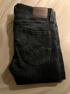 Mörkblå Replay jeans med bakficka - Säljer ett par mörkblå jeans från Replay med klassisk femficksdesign och snygga detaljer på bakfickan. Jeansen har en rak passform och är tillverkade i slitstarkt denimtyg. Märkespatch i läder baktill i midjan och diskreta röda sömmar vid fickan.