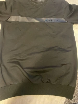 Svart Adidas topp med logga - Svart topp från Adidas med rund halsringning och en bred, blank rand över bröstet där loggan syns. Modellen har korta ärmar och ribbad nederkant. Perfekt för en sportig och avslappnad stil.