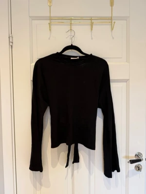 Nelly knyt topp - Trendig svart långärmad topp med knyt och öppen rygg. Perfekt för dig som gillar en stilren men ändå edgy look. Toppen har en croppad passform och är tillverkad i mjukt bomullsmaterial.