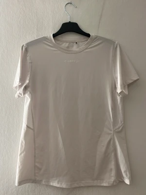 Vit tränings-tshirt från Craft - Snygg vit tränings-tshirt från Craft med rund hals och korta ärmar. Tillverkad i ett lätt och ventilerande funktionsmaterial som passar perfekt till löpning eller gym. Diskret logga framtill och meshpaneler på sidorna för extra andningsförmåga. Nästan aldrig använd 